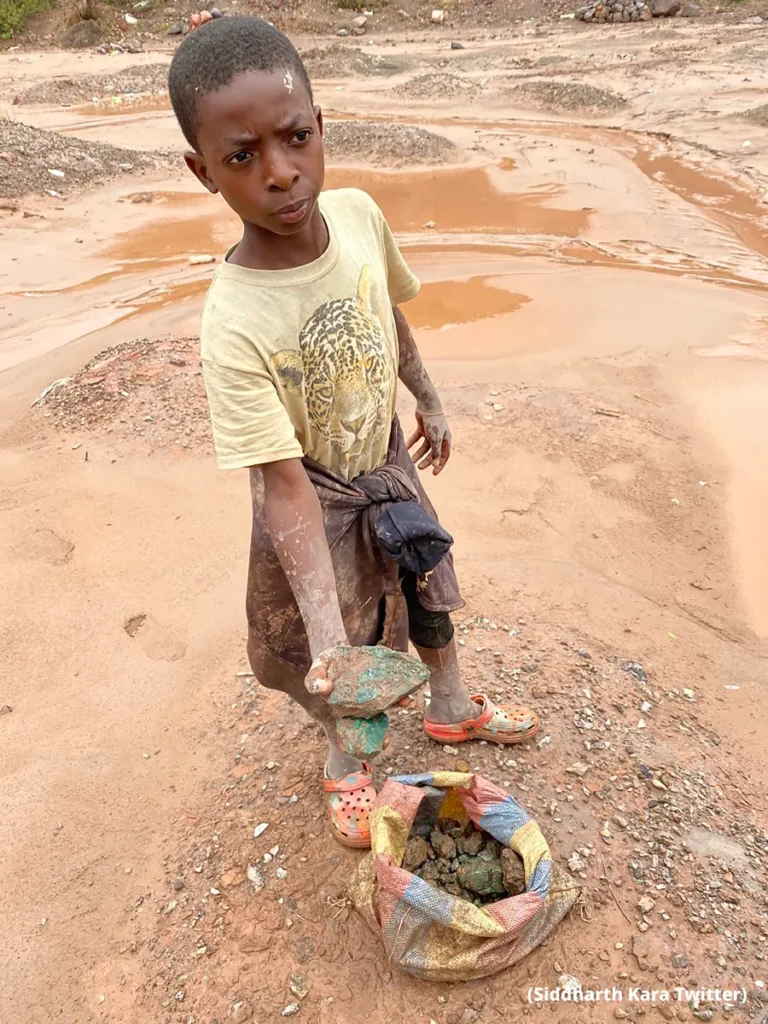 Young child holding green-stained rocks beside a sack at a muddy mining site